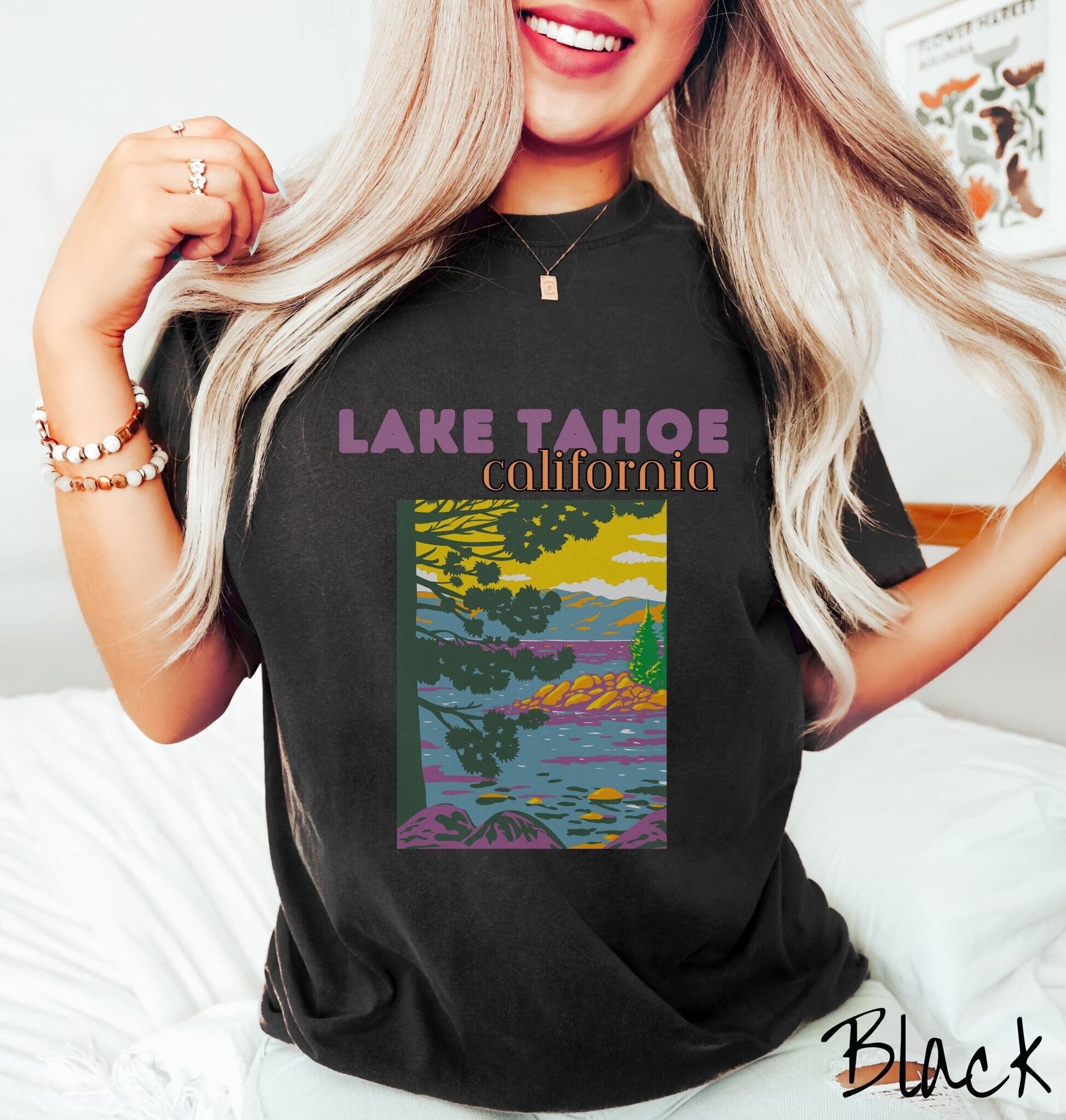 A woman wearing a vintage, black colored shirt with the text Lake Tahoe at the top in purple and orange font, respectively, and below is a beautiful picture of the ocean through the trees and rolling hills in the background.