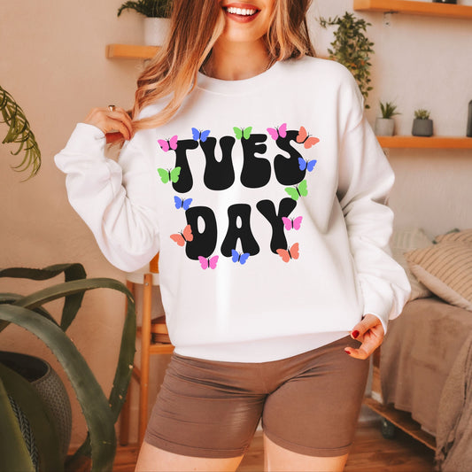 A woman wearing a vintage, white colored sweatshirt with the text centered on the front TUES then DAY below it, with pink, green, blue, and orange butterflies scattered amongst the letters.