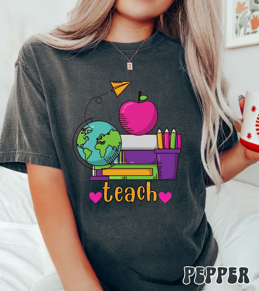 A woman wearing a vintage, pepper colored shirt with the word teach in between two pink hearts, and above are a stack of green, purple, and white books, a pink apple and a colorful pencil holder, a globe, and a yellow paper airplane flying above.