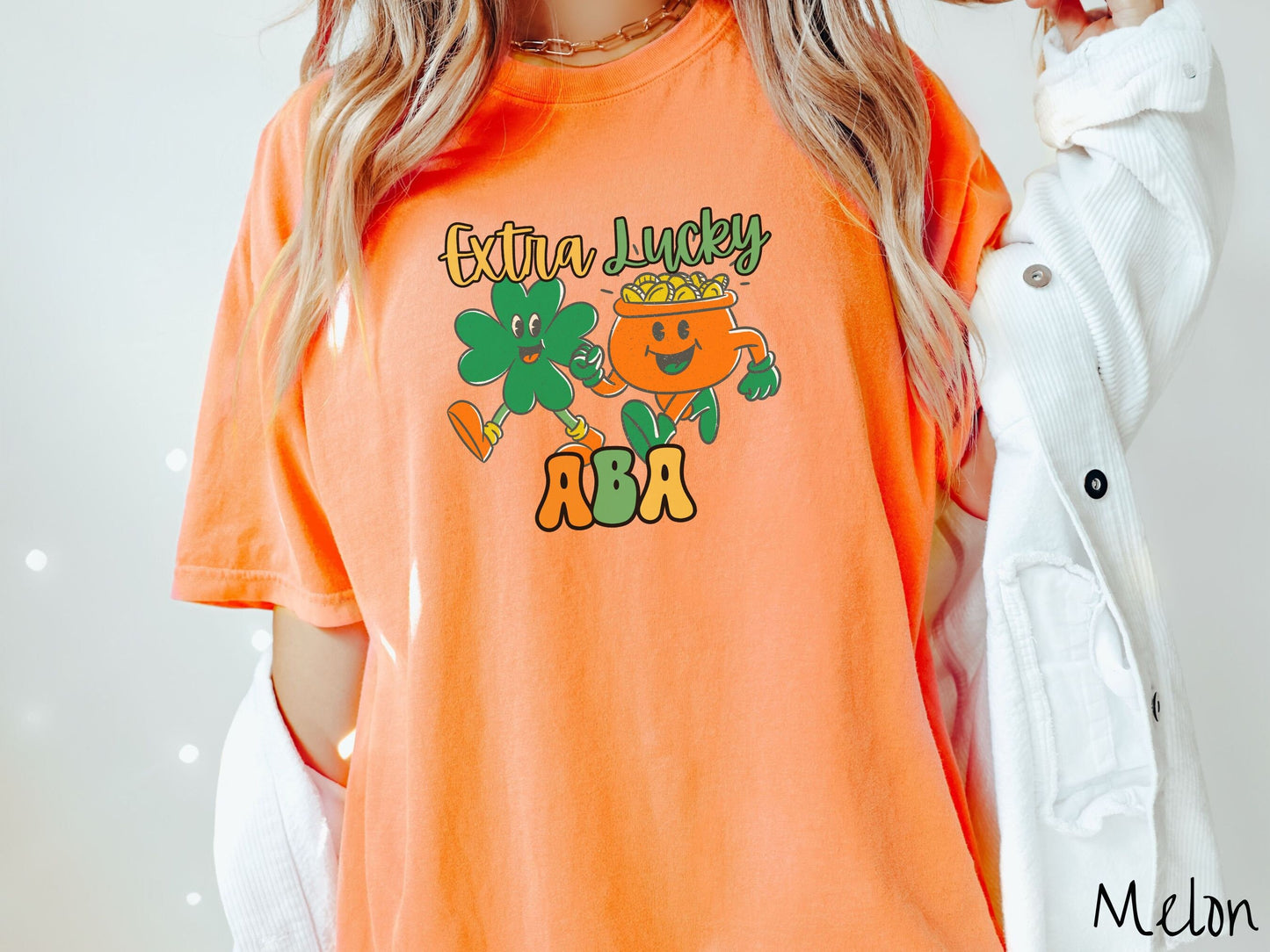 A woman wearing a vintage, melon colored shirt with the text Extra Lucky ABA in yellow, orange, and green font. Between the text are a green clover and orange pot of gold smiling and walking together in step.