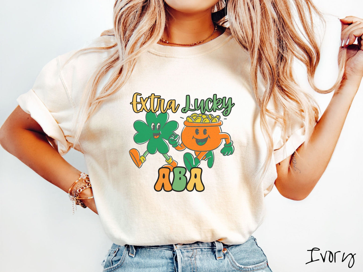 A woman wearing a vintage, ivory colored shirt with the text Extra Lucky ABA in yellow, orange, and green font. Between the text are a green clover and orange pot of gold smiling and walking together in step.