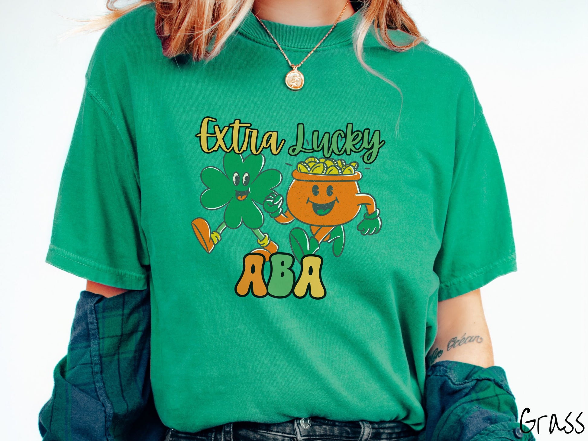 A woman wearing a vintage, grass colored shirt with the text Extra Lucky ABA in yellow, orange, and green font. Between the text are a green clover and orange pot of gold smiling and walking together in step.