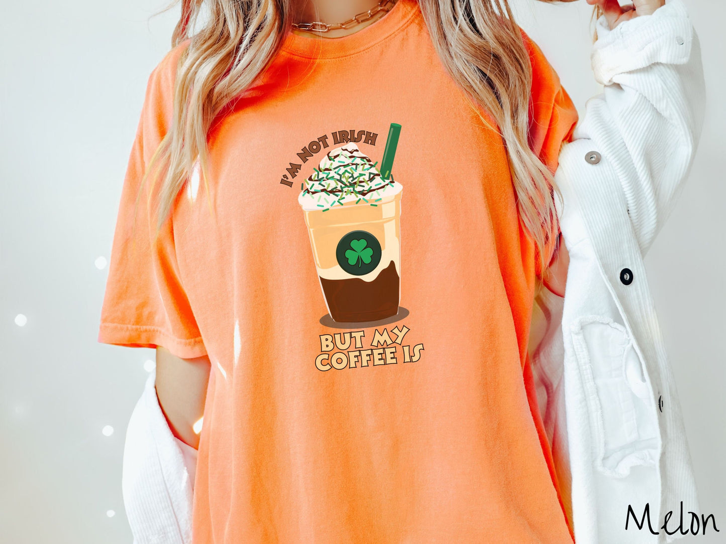 A woman wearing a vintage, melon colored shirt with the text Im not Irish but my coffee is in brown and yellow text, between the text is a green and yellow coffee cup with a green clover and  whipped cream and chocolate drizzle on top.