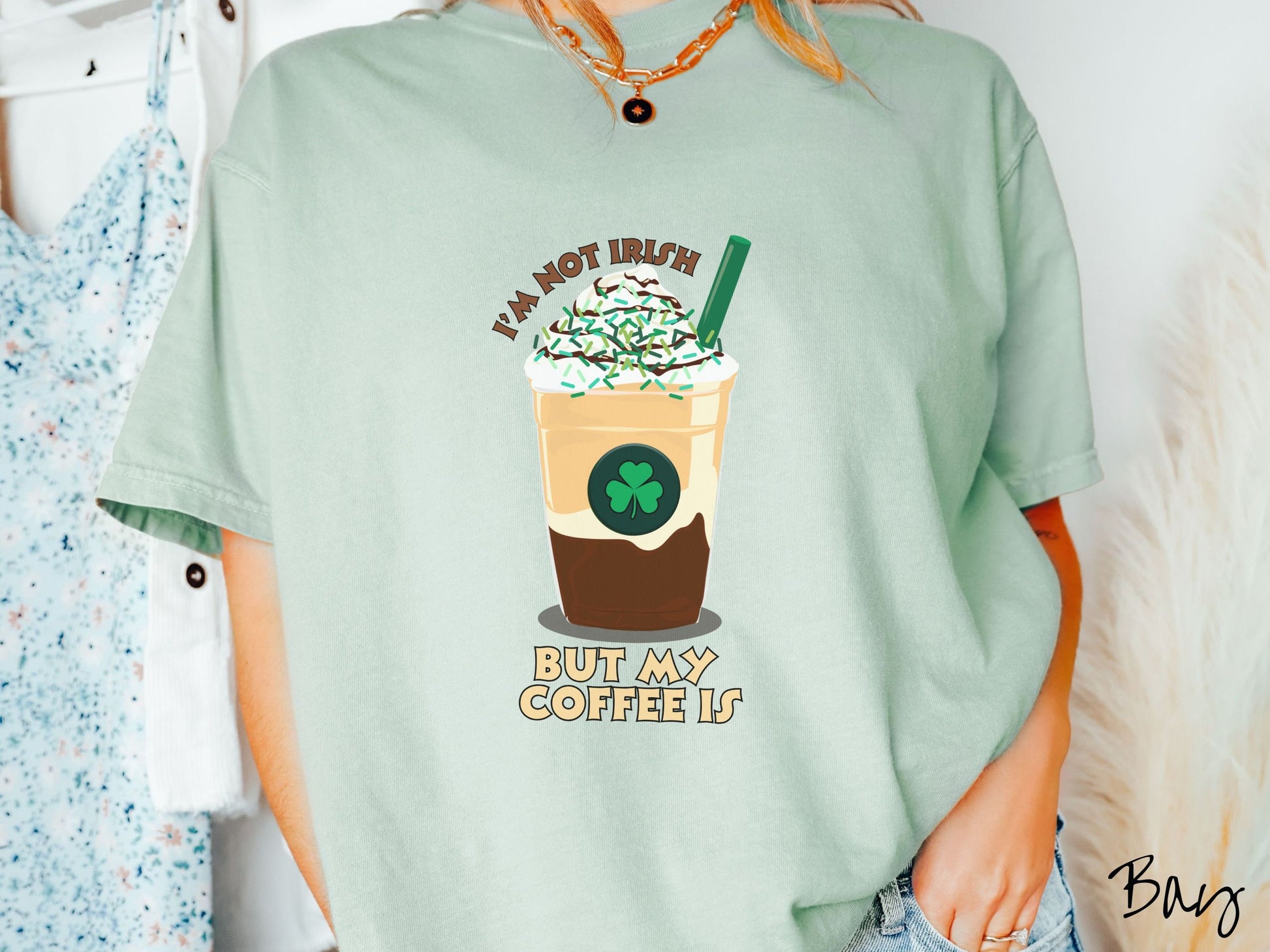 A woman wearing a vintage, bay colored shirt with the text Im not Irish but my coffee is in brown and yellow text, between the text is a green and yellow coffee cup with a green clover and  whipped cream and chocolate drizzle on top.