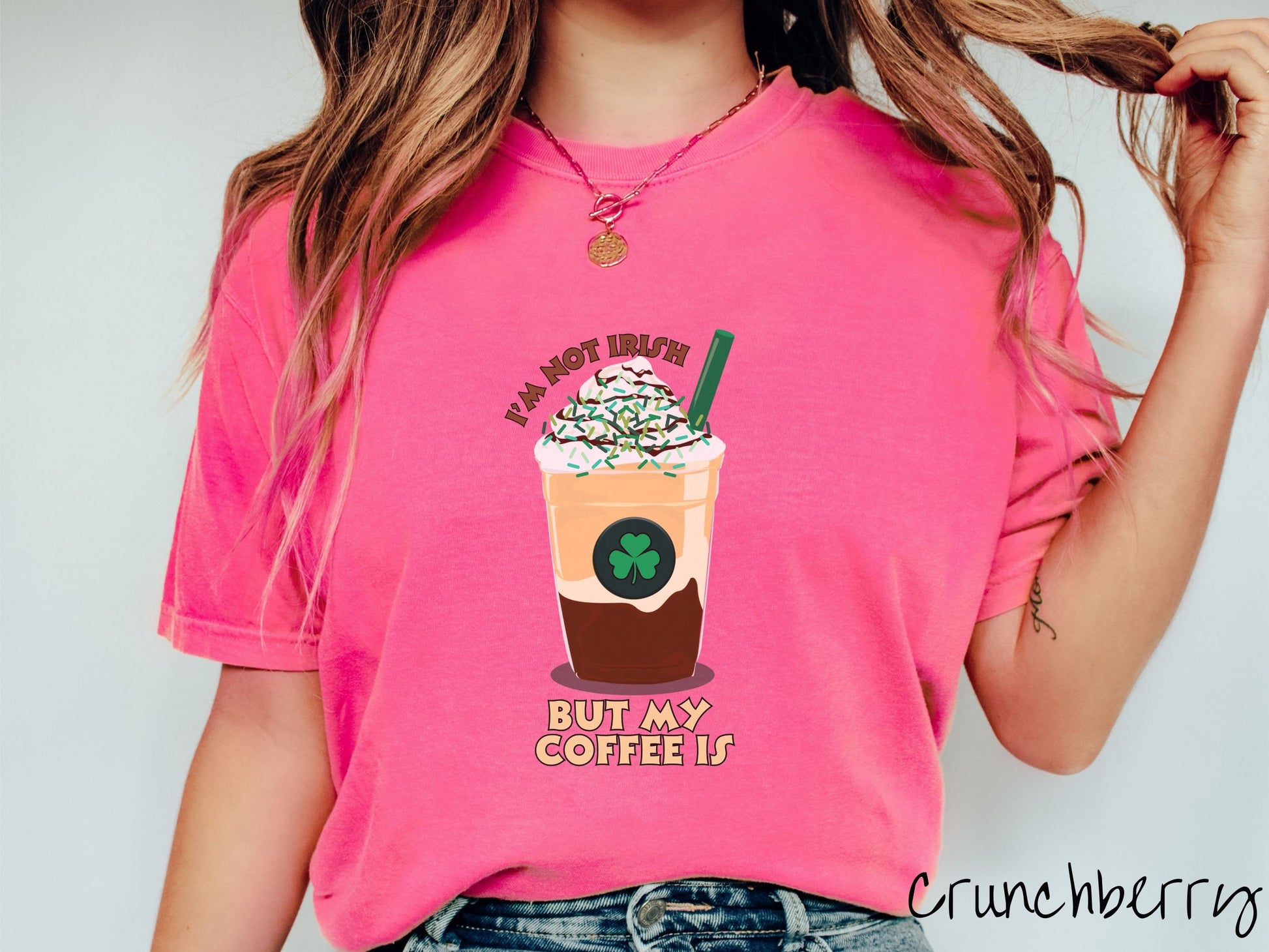 A woman wearing a vintage, crunchberry colored shirt with the text Im not Irish but my coffee is in brown and yellow text, between the text is a green and yellow coffee cup with a green clover and  whipped cream and chocolate drizzle on top.