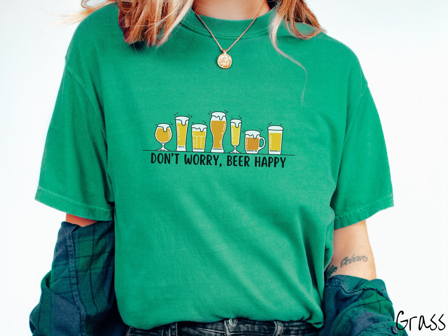 A woman wearing a vintage, grass colored shirt with the text Dont Worry, Beer happy, and above this are varying sizes and shapes of beer glasses with yellow and orange ber all of them have foam coming out the tops.