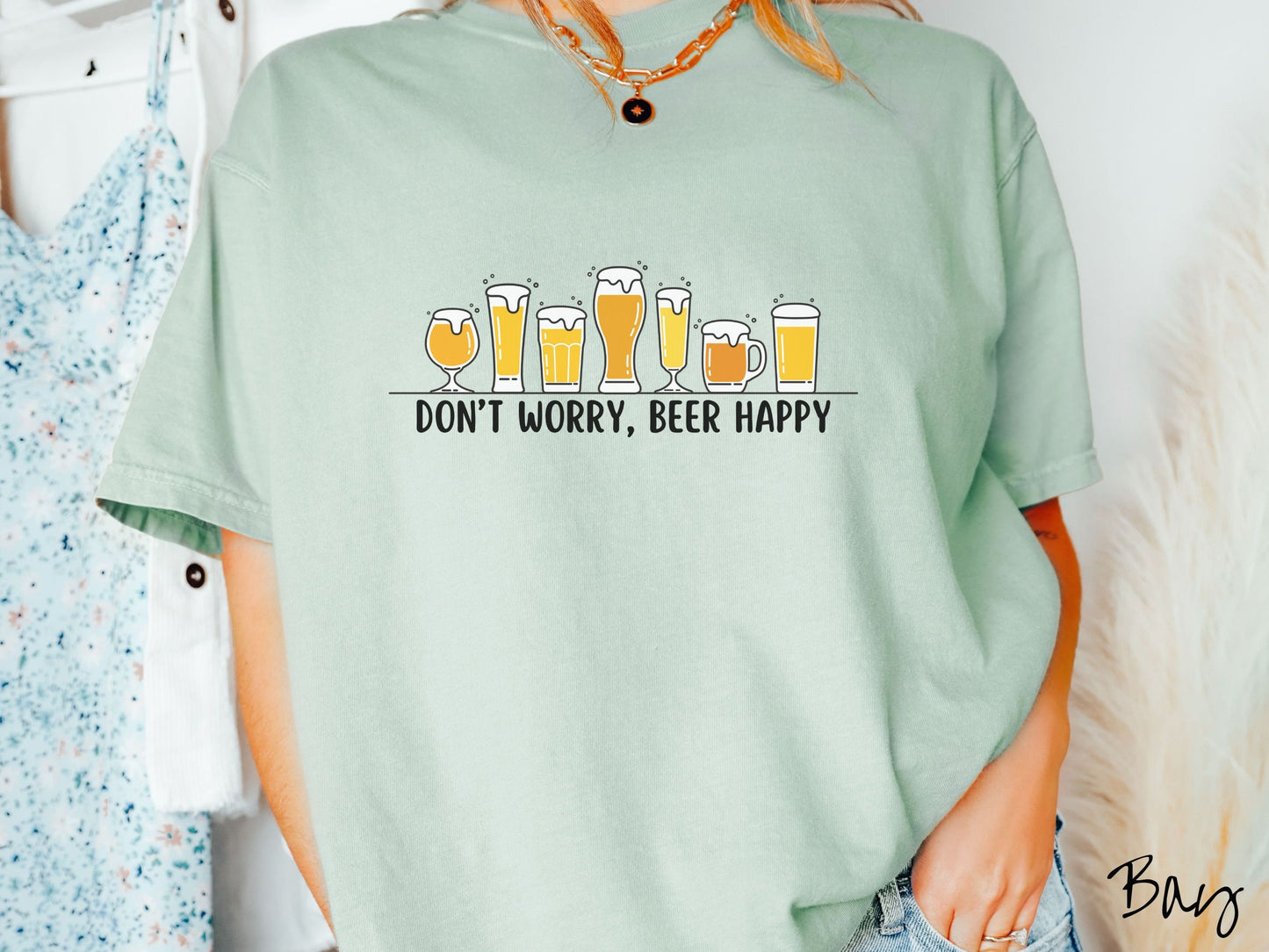 A woman wearing a vintage, bay colored shirt with the text Dont Worry, Beer happy, and above this are varying sizes and shapes of beer glasses with yellow and orange ber all of them have foam coming out the tops.