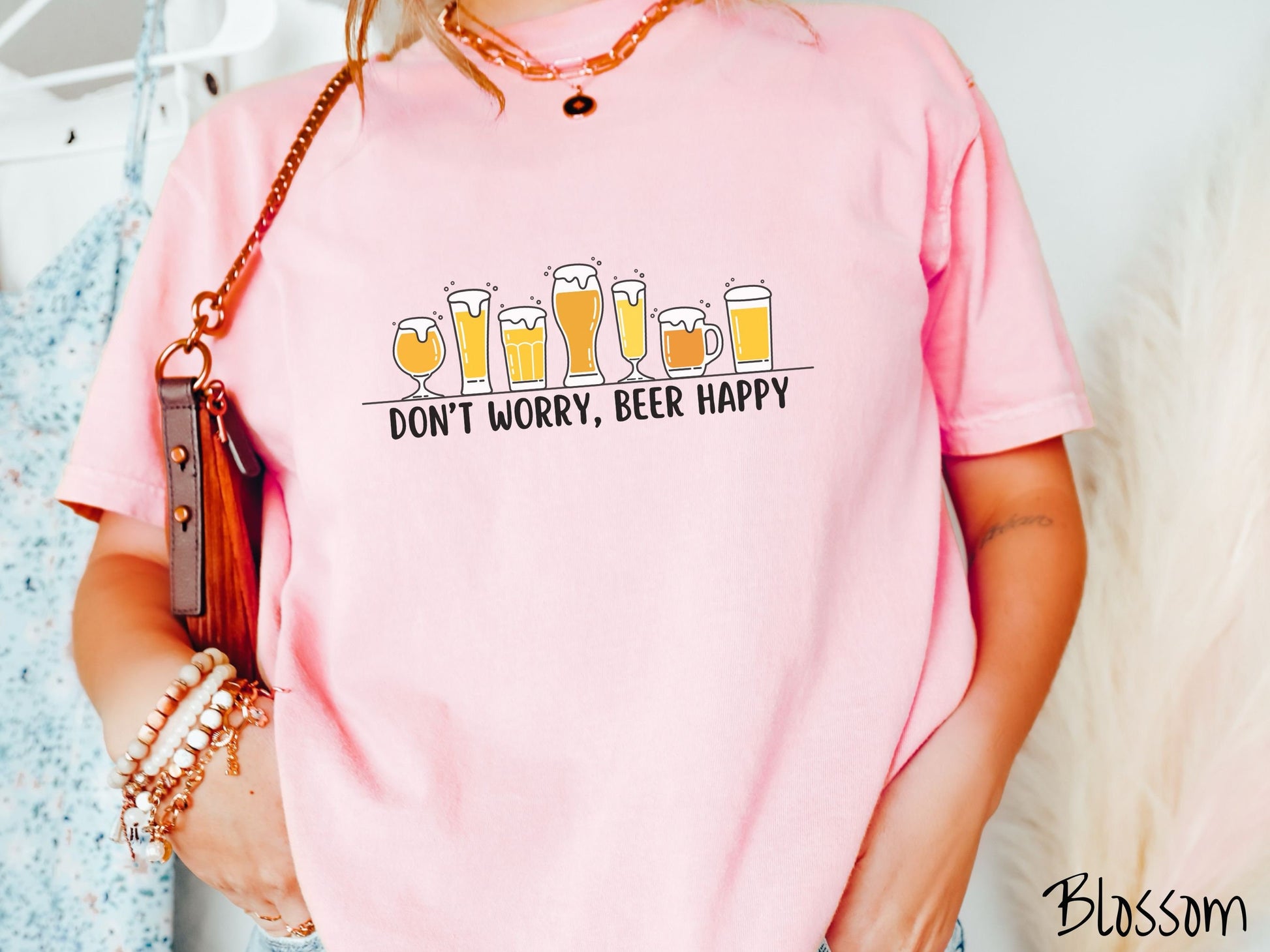 A woman wearing a vintage, blossom colored shirt with the text Dont Worry, Beer happy, and above this are varying sizes and shapes of beer glasses with yellow and orange ber all of them have foam coming out the tops.