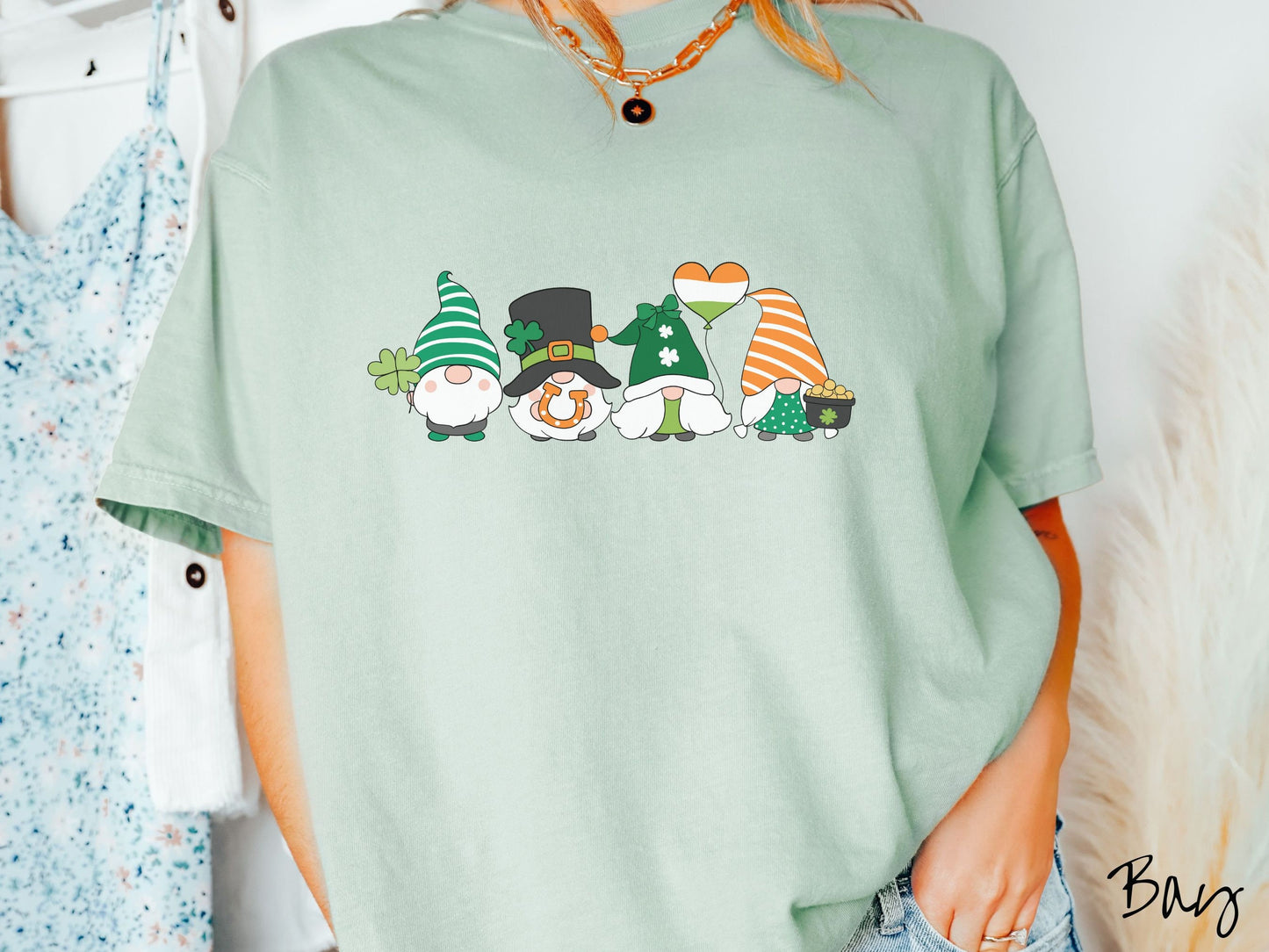 A woman wearing a vintage, bay colored shirt with four festive gnomes wearing varying green, white, and orange hats holding a green clover, an inflatable balloon, an orange horse shoe, and a pot of gold.