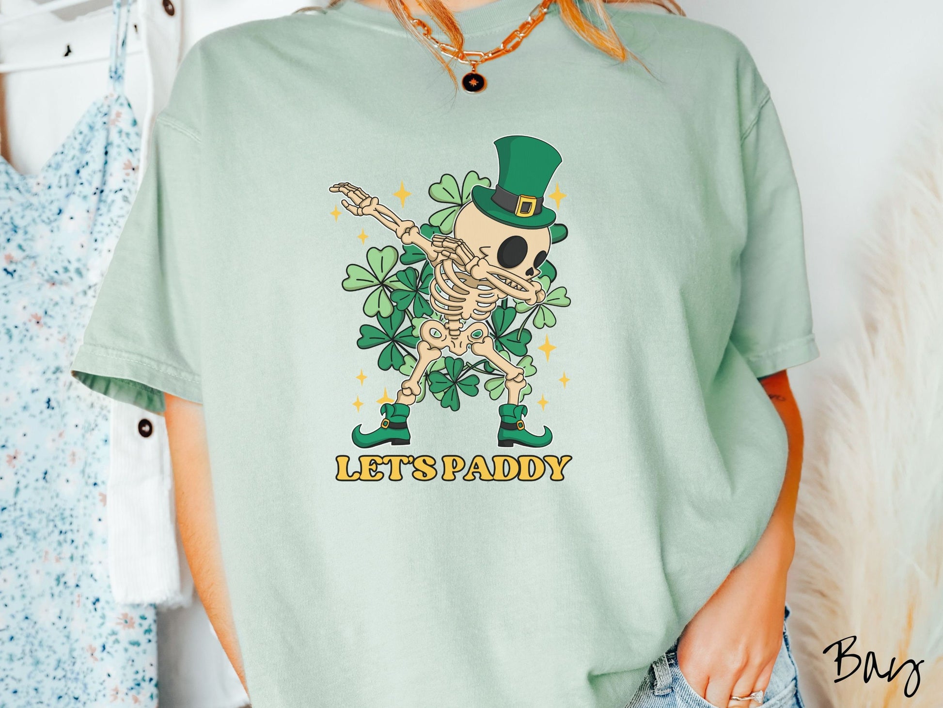 A woman wearing a vintage, bay colored shirt with the text Lets Paddy and below that is a leprechaun wearing a green top hat and green elf shoes doing the dab dance and there are green clovers and golden stars in the background.