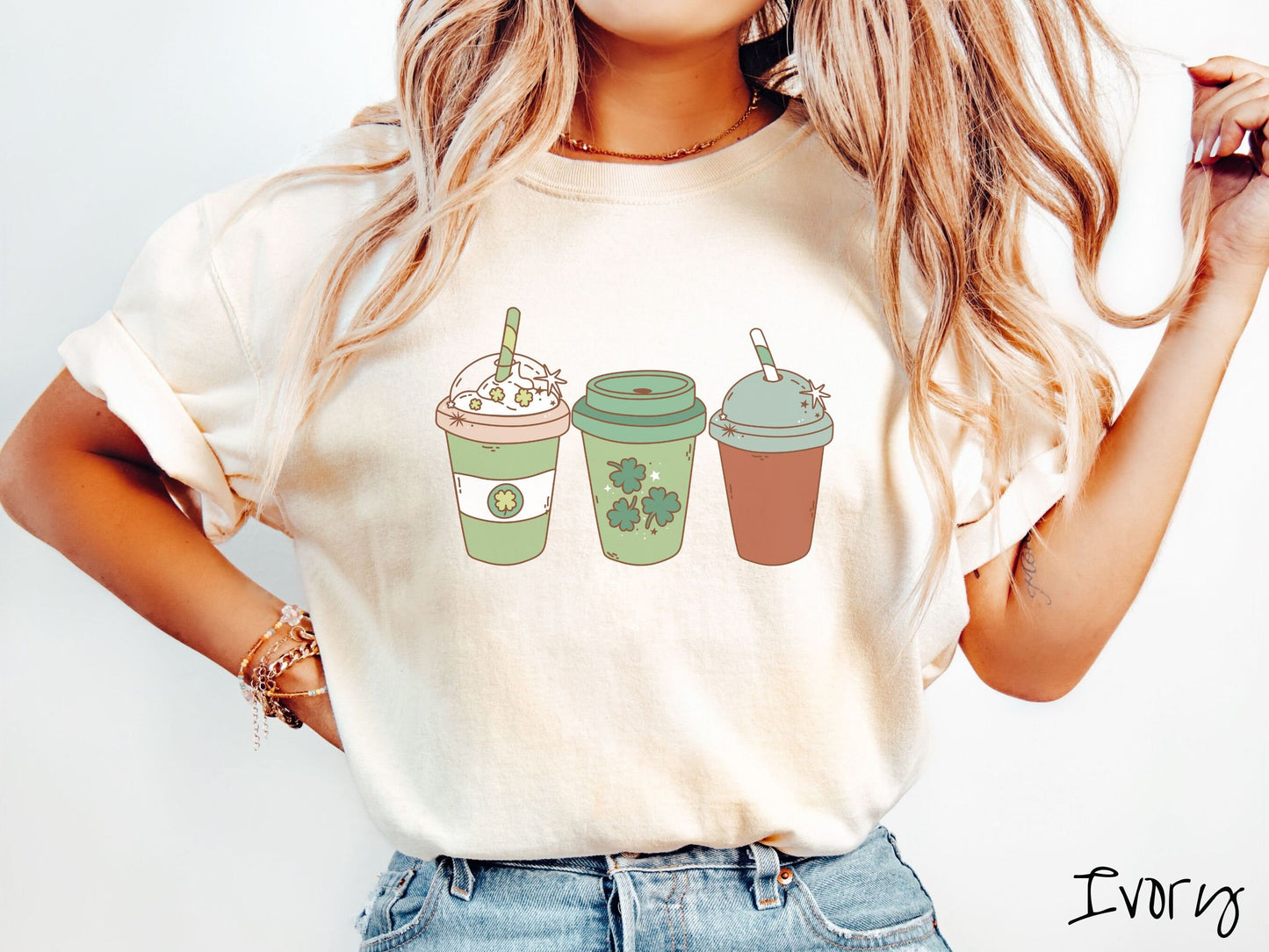 A woman wearing a vintage, ivory colored shirt with three coffee cups, one is white and green with a clear lid showing whipped cream topping with green clover sprinkles, another green cup with green clovers, and the third is brown with a green lid.