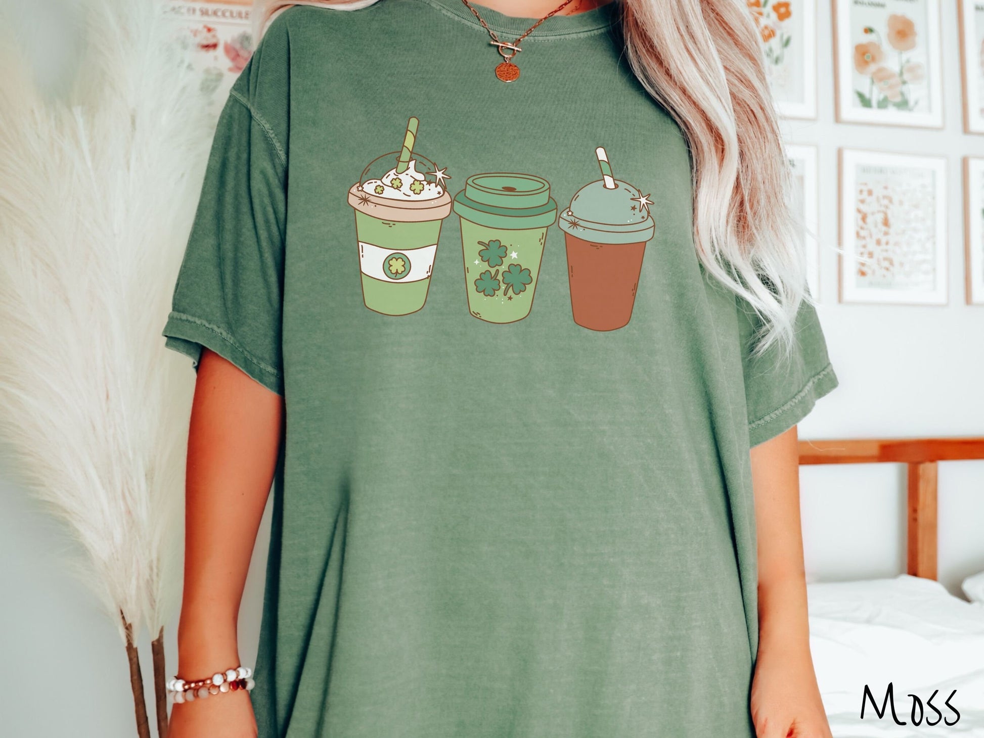 A woman wearing a vintage, moss colored shirt with three coffee cups, one is white and green with a clear lid showing whipped cream topping with green clover sprinkles, another green cup with green clovers, and the third is brown with a green lid.