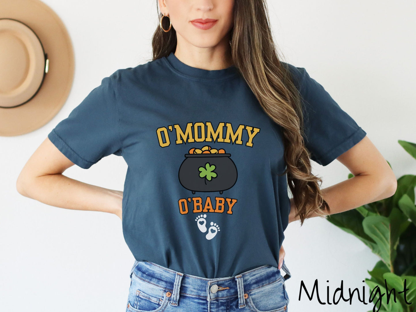 A woman wearing a vintage, midnight colored shirt with text O Mommy O Baby in yellow and orange font, a pot of gold with a green clover on it and yellow and orange gold coins inside. Below all this are two white baby footprints with pink hearts.
