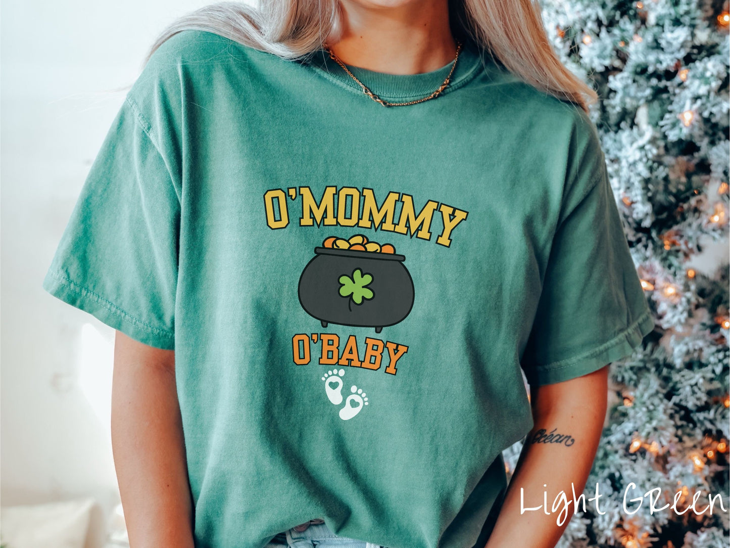 A woman wearing a vintage, light green colored shirt with text O Mommy O Baby in yellow and orange font, a pot of gold with a green clover on it and yellow and orange gold coins inside. Below all this are two white baby footprints with pink hearts.