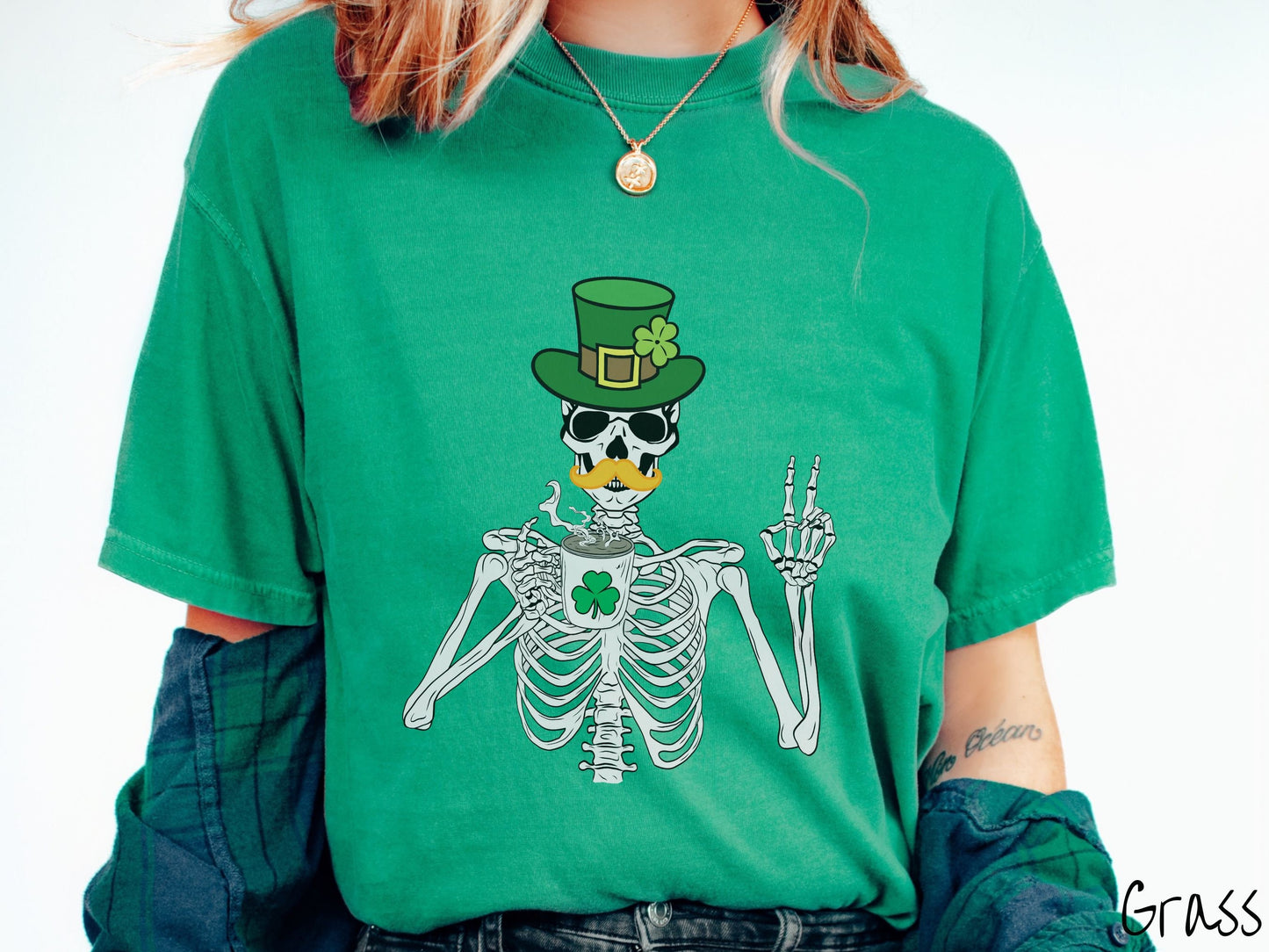 A woman wearing a vintage, grass colored shirt with a skeleton wearing a green top hat with a green clover and an orange mustache showing the peace sign with its left hand and drinking out of a steaming hot coffee cup with a three leaf clover on it.
