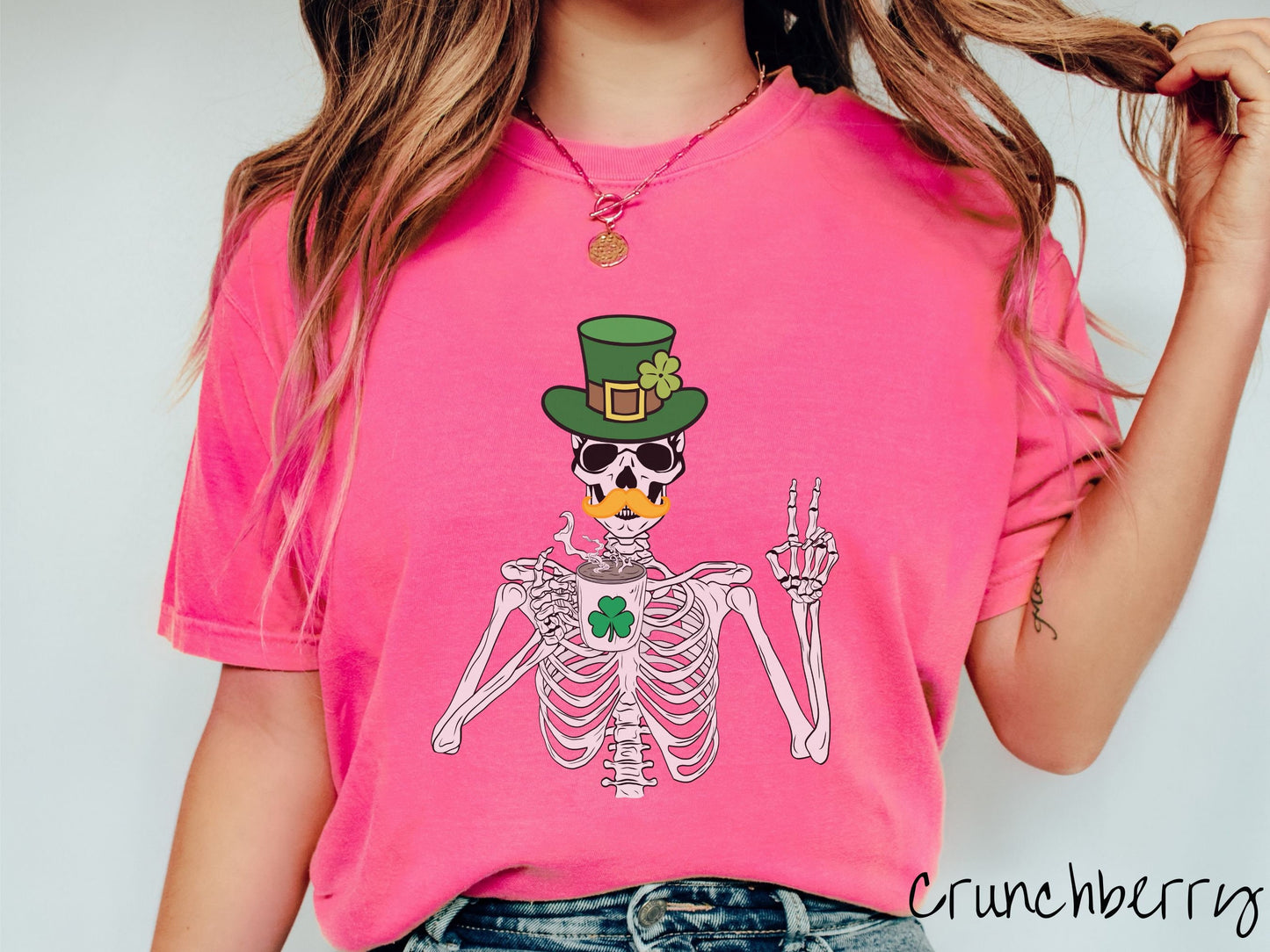 A woman wearing a vintage, crunchberry colored shirt with a skeleton wearing a green top hat and an orange mustache showing the peace sign with its left hand and drinking out of a steaming hot coffee cup with a three leaf clover on it.