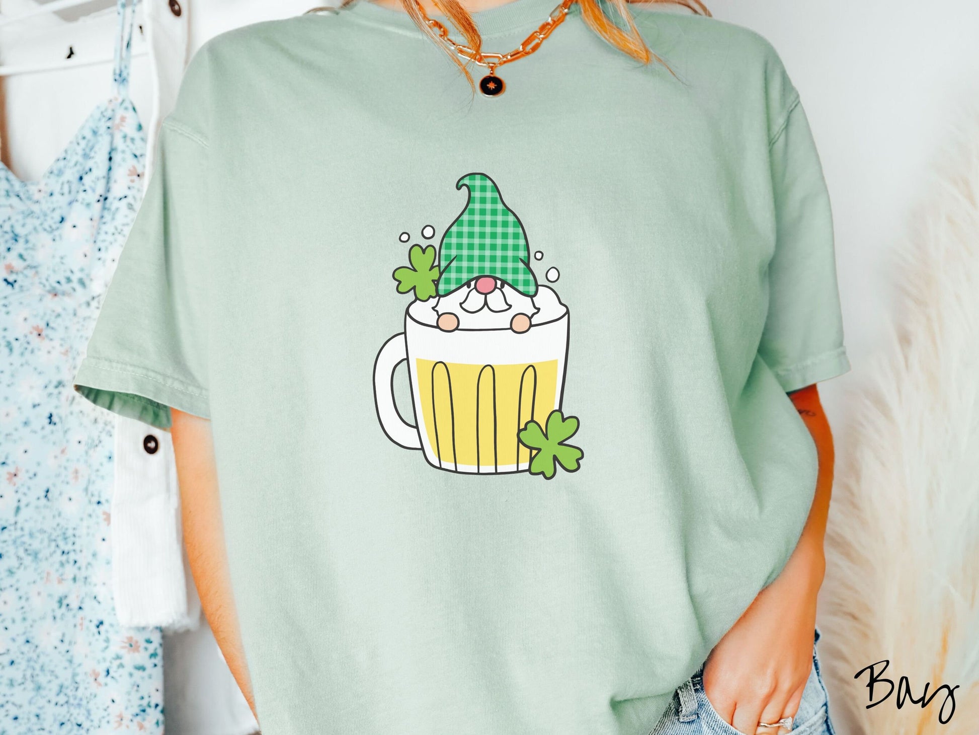 A woman wearing a vintage, bay colored shirt with a beer filled mug in the center with a green hat wearing gnome peeking over the top of the glass and green clovers in the background.