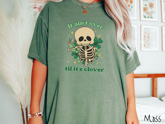 A woman wearing a vintage, moss colored shirt with the text It aint over til its clover and in between the text is a smiling skeleton holding a peace sign with its right hand, with many green three leaf clovers and golden stars in the background.