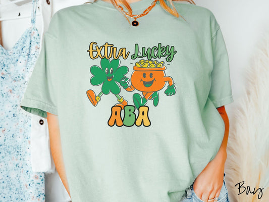 A woman wearing a vintage, bay colored shirt with the text Extra Lucky ABA in yellow, orange, and green font. Between the text are a green clover and orange pot of gold smiling and walking together in step.