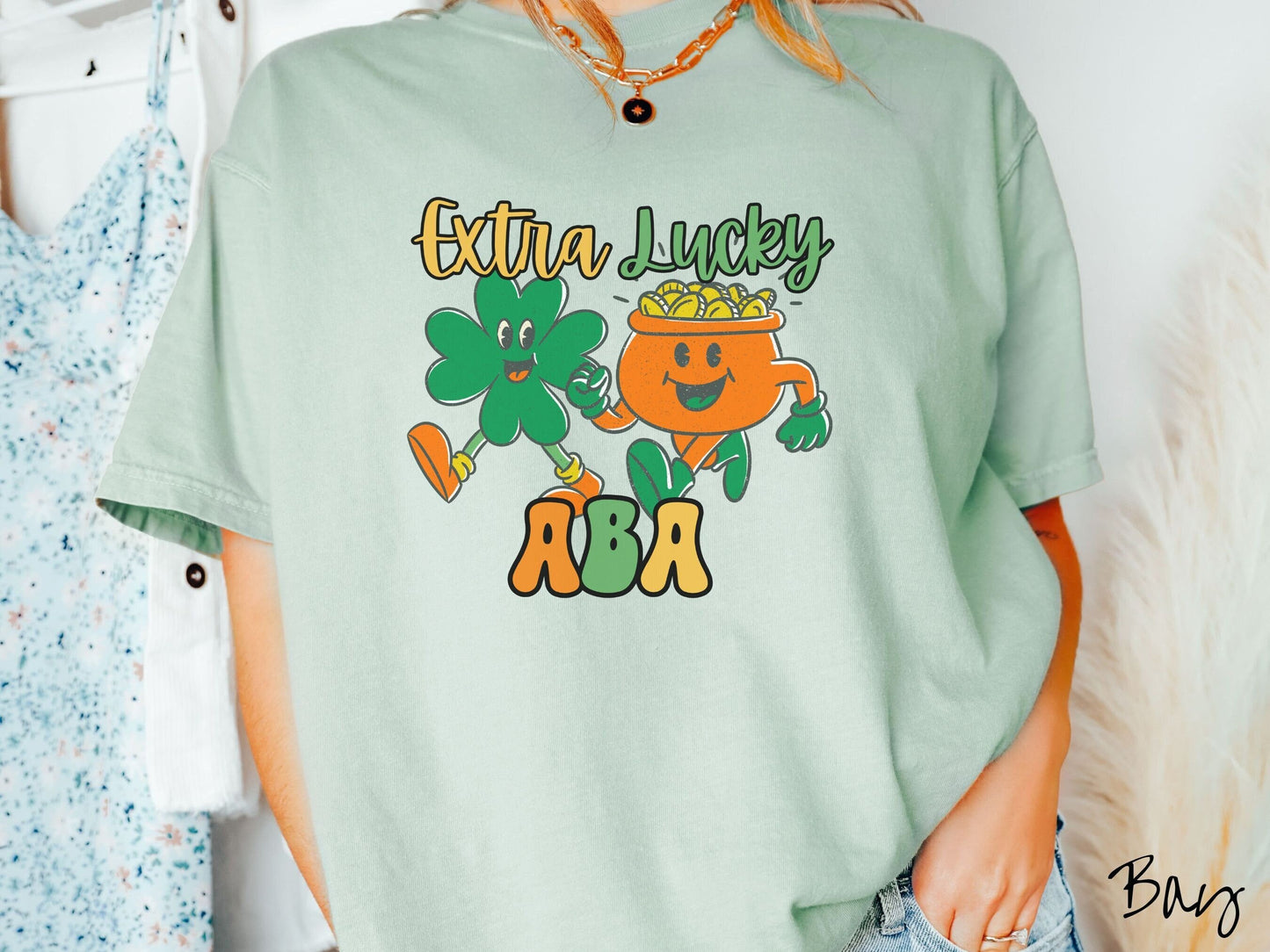 A woman wearing a vintage, bay colored shirt with the text Extra Lucky ABA in yellow, orange, and green font. Between the text are a green clover and orange pot of gold smiling and walking together in step.