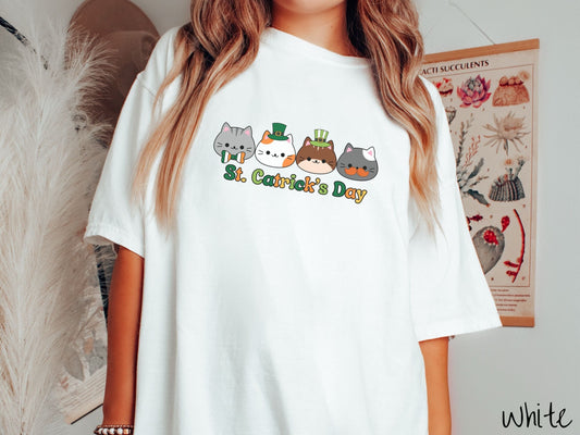 A woman wearing a vintage, white colored shirt with four different cats lined across the front, one with a green, white, and orange bowtie, two with green top hats, and the fourth with an orange mustache, and below is the saying St. Catricks Day.