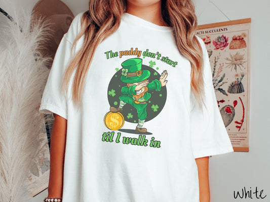 A woman wearing a vintage, white colored shirt with the text The paddy don&#39;t start til I walk in and below that is a leprechaun doing the dab dance with his right foot resting on a golden coin and green three leaf clovers in the background.