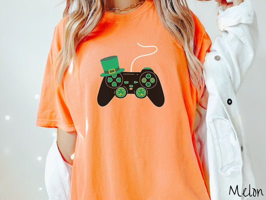 A woman wearing a vintage, melon colored shirt with a black video game controller with green buttons and joysticks, the analog control sticks are in the shape of green leaf clovers, and there is a green top hat hanging off the top left trigger.