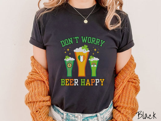 A woman wearing a vintage, black colored shirt with text Dont Worry Beer Happy in green, yellow, orange, and white font, in between the text are three beer mugs with Irish-themed clovers, rainbows, and hearts on them.