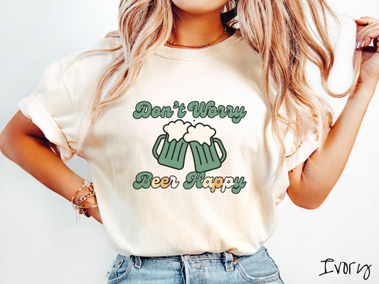 A woman wearing a vintage, ivory colored shirt with the text Dont Worry Beer Happy in green, yellow, white, and orange font, in between the text are two green beer glasses with beer foaming over the tops clinking together cheering.