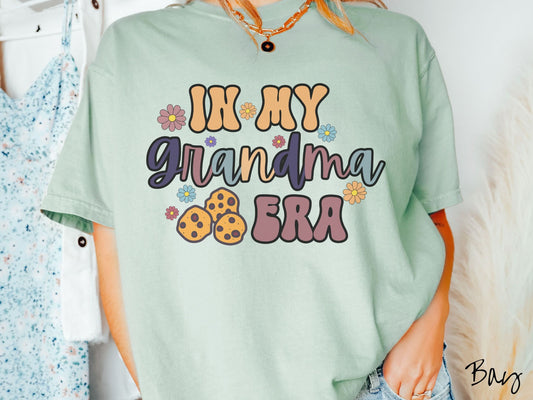 A woman wearing a cute light green shirt with the text saying In My Grandma era with flowers and chocolate chip cookies mixed within the text.