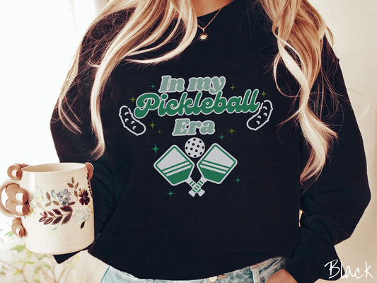 A woman wearing a cute black colored sweatshirt with the text In My Pickleball Era across the top, and below are 2 pickleball paddles and a ball. There are pickles at the bottom left and right corners of the text.