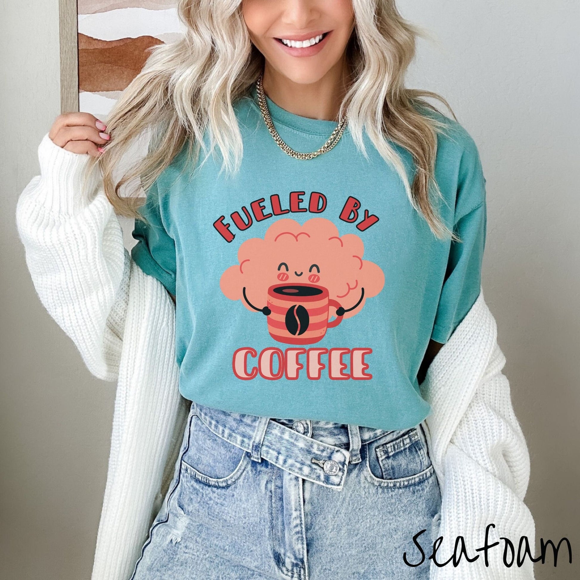 A woman wearing a cute seafoam colored shirt with the text Fueled by Coffee and a puffy cloud smiling holding a cup of coffee.