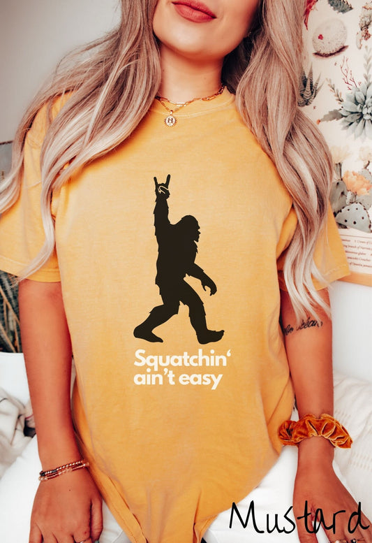 A woman wearing a cute mustard colored shirt with text saying Squatchin aint easy and a silhouette of Bigfoot holding up the rock on hand sign.