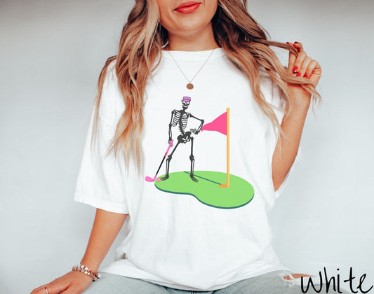 A woman wearing a cute white colored shirt with a black skeleton posing on the golfing green with a pink putter, pink golf ball, and a pink flag in the hole.