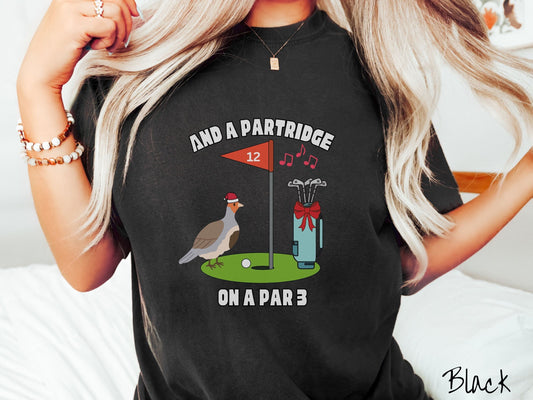 A woman wearing a cute black colored shirt with a partridge bird on a golf green next to a hole with a flag in it with #12 on it. There is also a set of golf clubs with a red bow and music playing in the air.