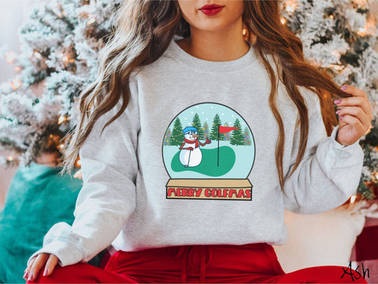 A woman wearing a cute ash colored sweatshirt with red text saying Merry Golfmas and a picture of a snowman giving the thumbs up on the golf green next to the hole with a flag in it.