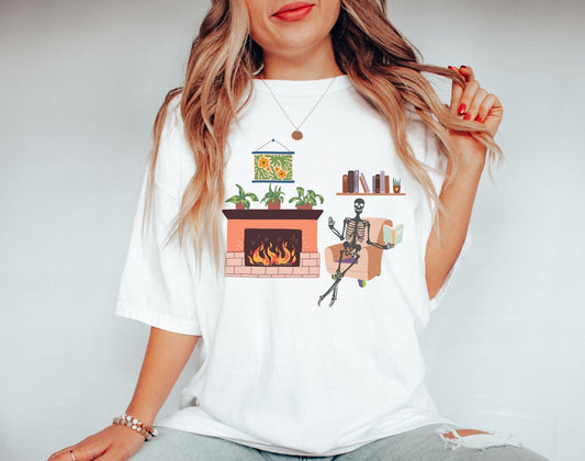 A woman wearing a cute white colored shirt with a picture of a skeleton sitting in a cushy chair reading a book under a bookshelf, a fireplace with a roaring fire to the right of the chair with plants on top of the hearth.