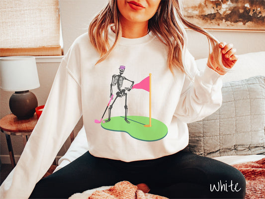 A woman wearing a cute white colored sweatshirt with a black skeleton posing on the golfing green with a pink putter, pink golf ball, and a pink flag in the hole.