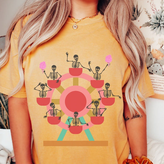 A woman wearing a cute mustard colored shirt with a ferris wheel filled with black skeletons waving and holding cotton candy as they go around the ride.