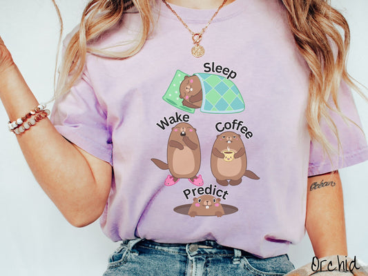 A woman wearing a cute orchid colored shirt with the text Sleep, Wake, Coffee, and Predict accompanied with pictures of a groundhog sleeping, a groundhog waking up yawning, a groundhog holding a steaming cup of coffee, and another exiting its hole.