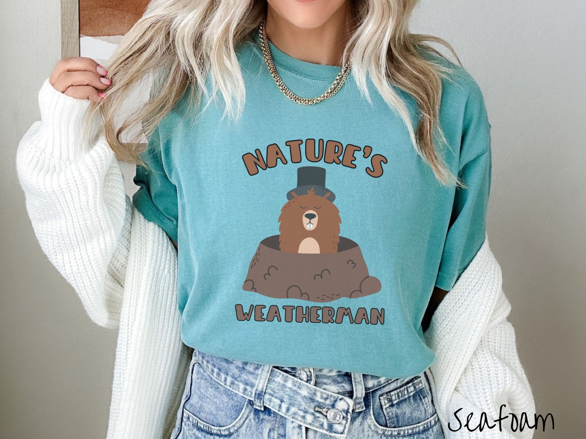 A woman wearing a cute seafoam colored shirt with the text Natures Weatherman sandwiching Phil the Punxsutawney PA groundhog who is wearing a black top hat and is coming out of his hole for Groundhogs Day.