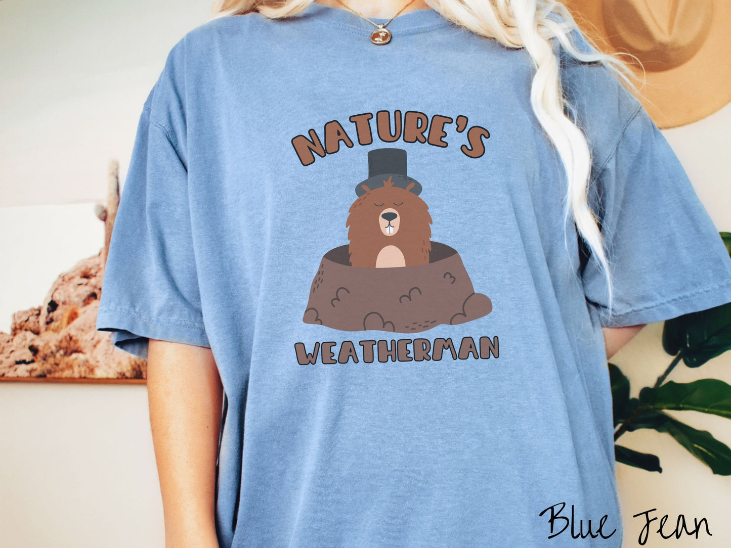 A woman wearing a cute blue jean colored shirt with the text Natures Weatherman sandwiching Phil the Punxsutawney PA groundhog who is wearing a black top hat and is coming out of his hole for Groundhogs Day.