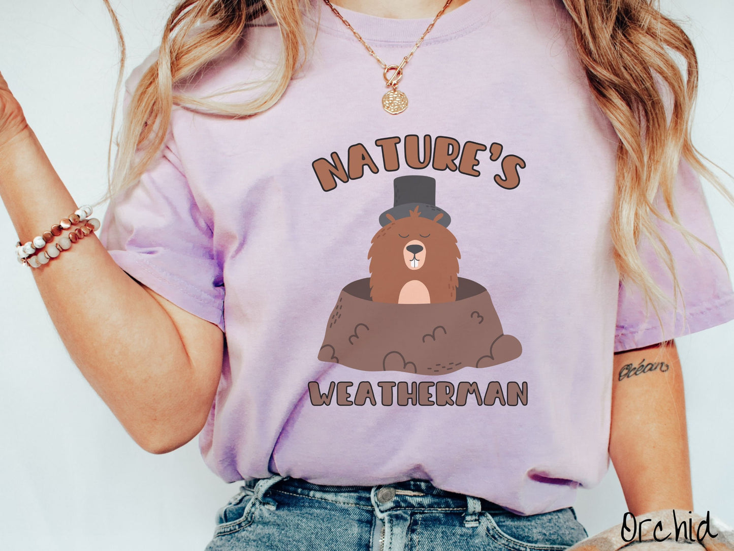 A woman wearing a cute orchid colored shirt with the text Natures Weatherman sandwiching Phil the Punxsutawney PA groundhog who is wearing a black top hat and is coming out of his hole for Groundhogs Day.