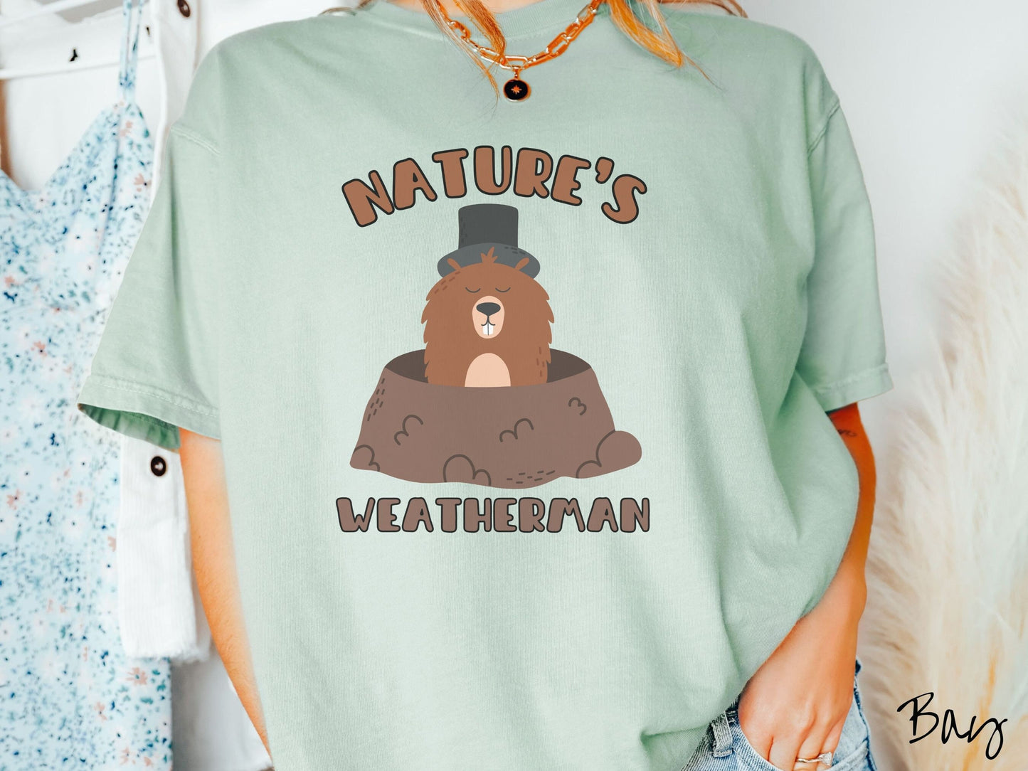 A woman wearing a cute bay colored shirt with the text Natures Weatherman sandwiching Phil the Punxsutawney PA groundhog who is wearing a black top hat and is coming out of his hole for Groundhogs Day.