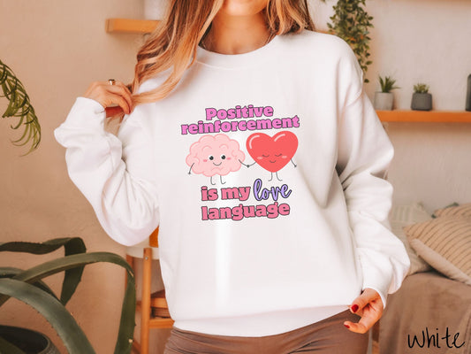 A woman wearing a cute, vintage white colored sweatshirt with the text Positive reinforcement is my love language in pink, purple, and red with a pink smiling brain holding hands with a red smiling heart.