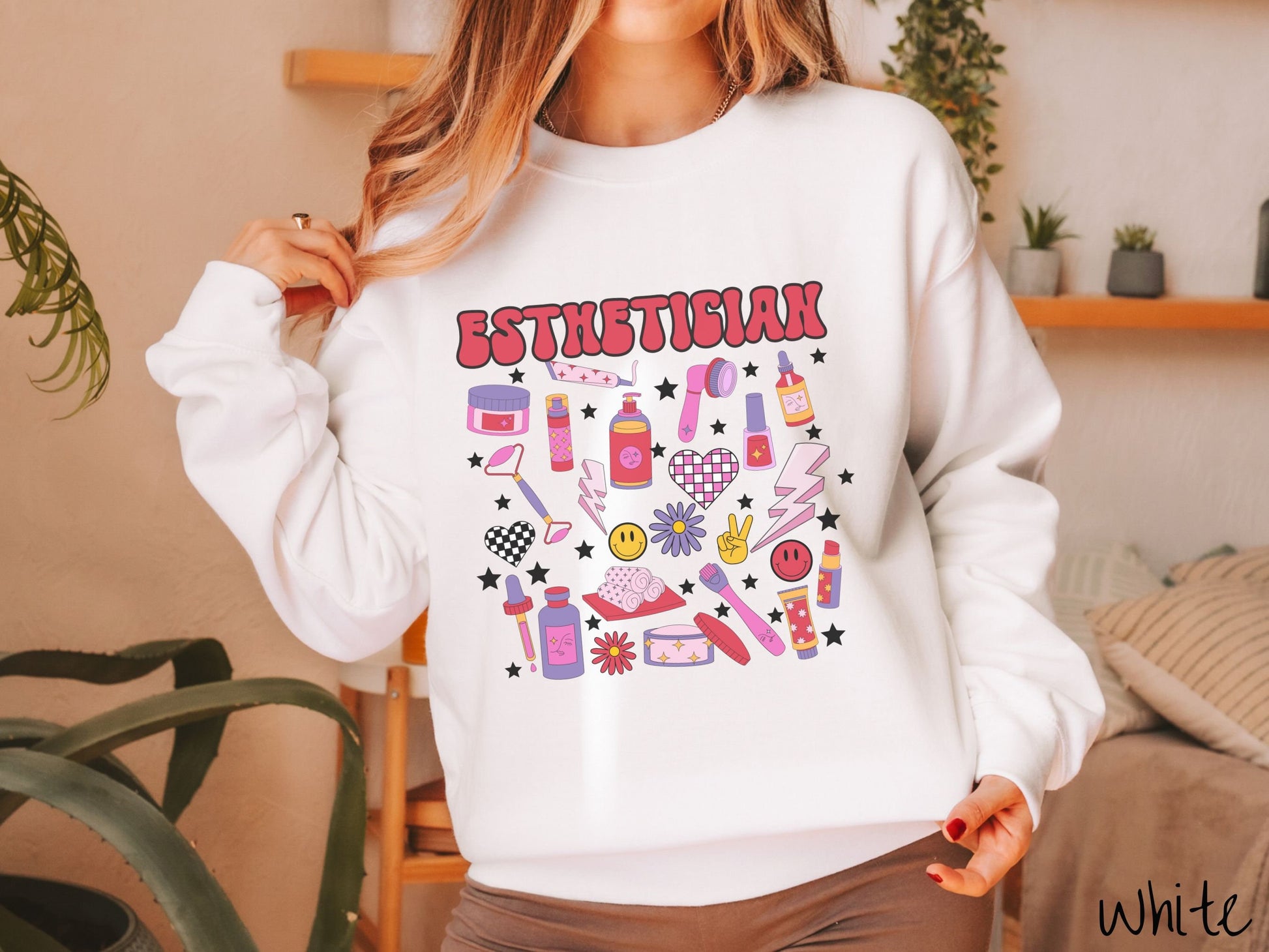 A woman wearing a cute white colored sweatshirt with the word Esthetician across the top and hair care items below like cream, rollers, hair care products, and droppers. Smiley faces, stars, and lightning bolts are spread out among the items.