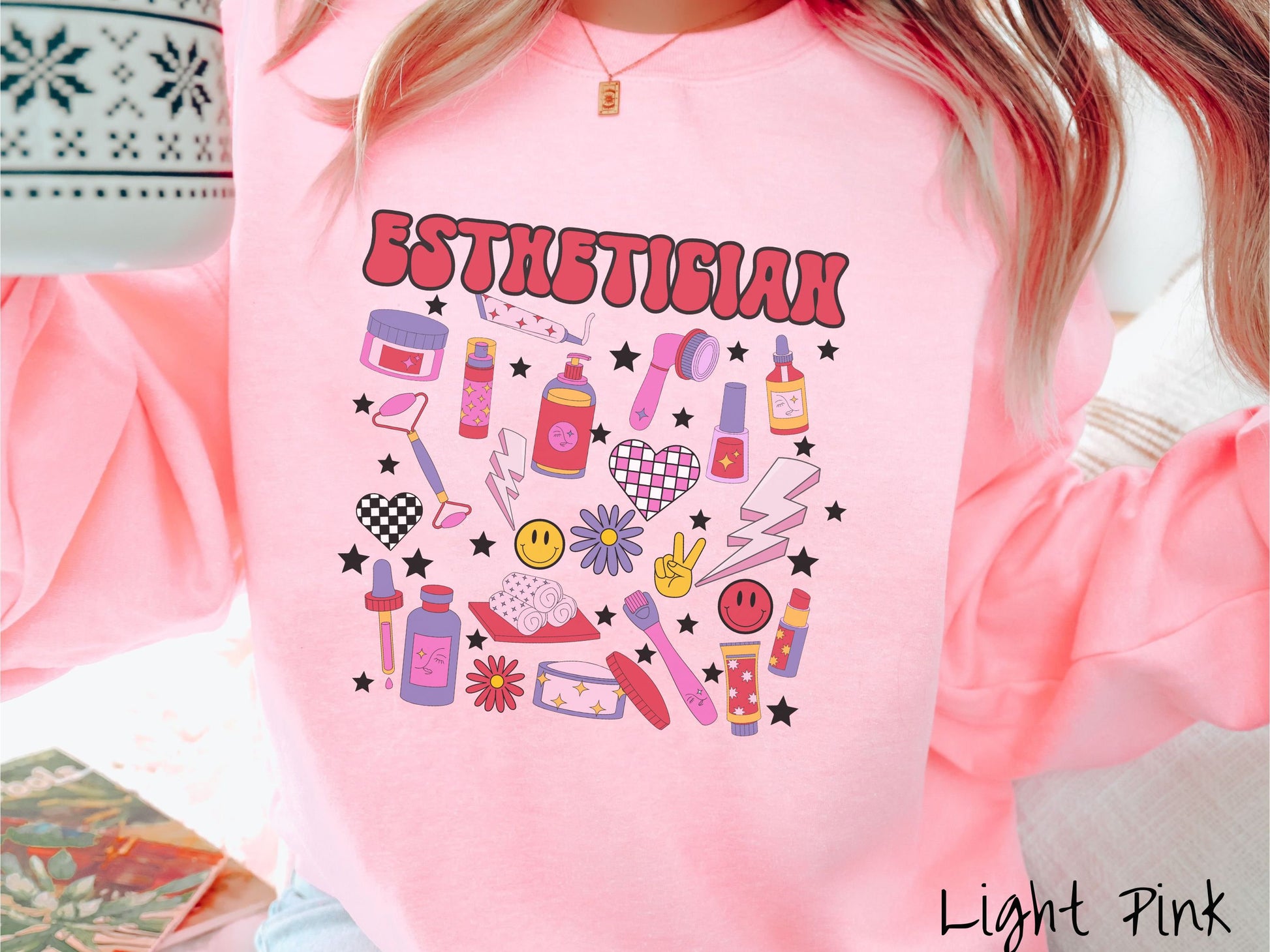 A woman wearing a cute light pink colored sweatshirt with the word Esthetician across the top and hair care items below like cream, rollers, hair care products, and droppers. Smiley faces, stars, and lightning bolts are spread out among the items.