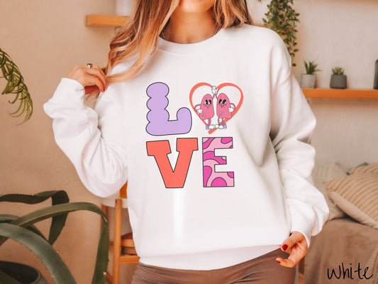 A woman wearing a cute white colored sweatshirt with the word LOVE spelled out with the L as a purple lung, the O as a heart with two lung figures inside high fiving, the V is orange, and the E is two shades of pink.