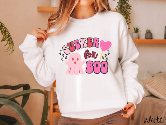 A woman wearing a cute white colored sweatshirt with the text saying Sucker for Boo, with a pink ghost with its arms open, a lollipop with a pink heart, and hearts floating in the air.