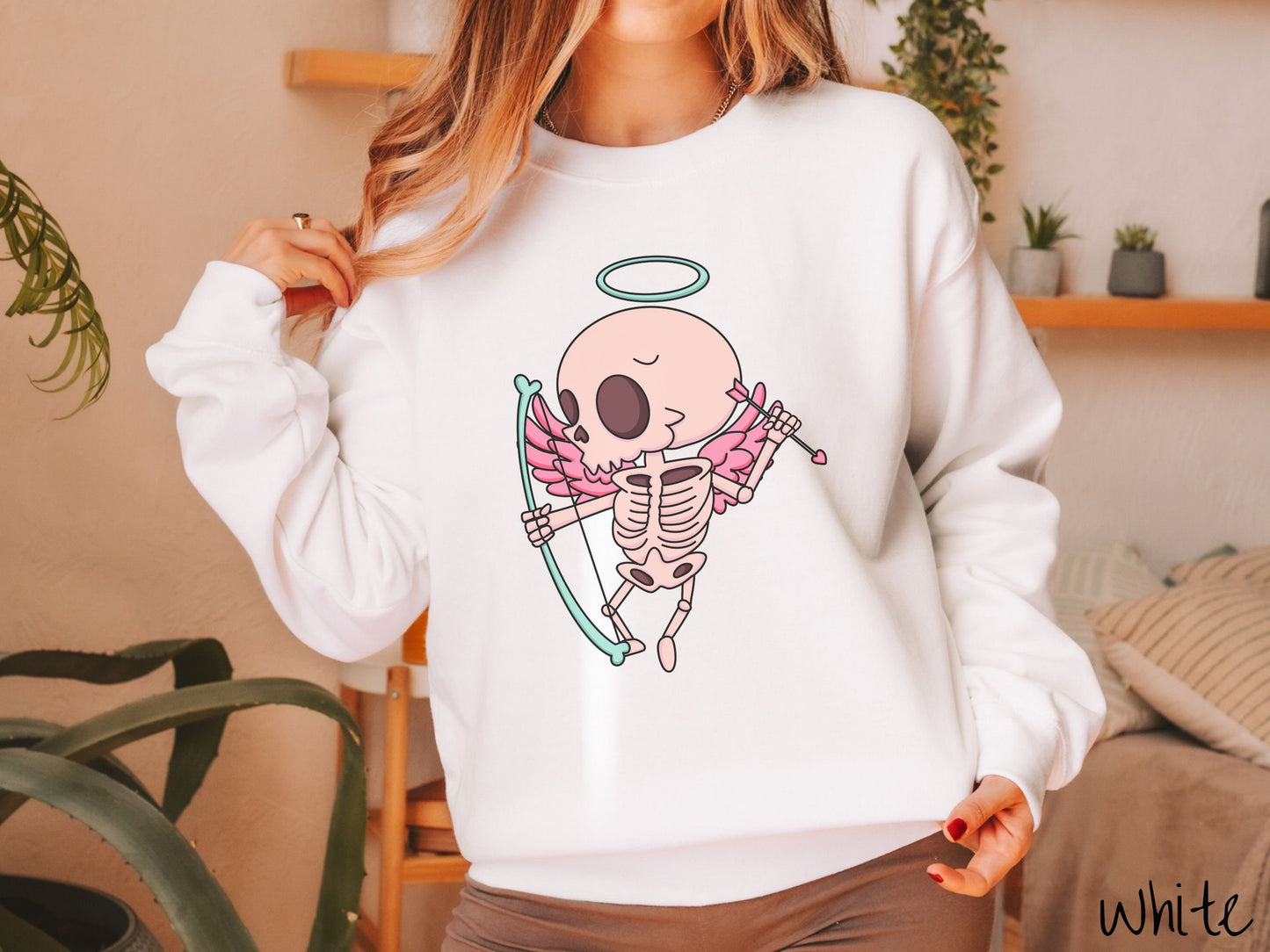 A woman wearing a cute white colored sweatshirt with a pink winged baby skeleton cupid with a halo holding a heart-tipped arrow in its left hand and a bow in its right hand.